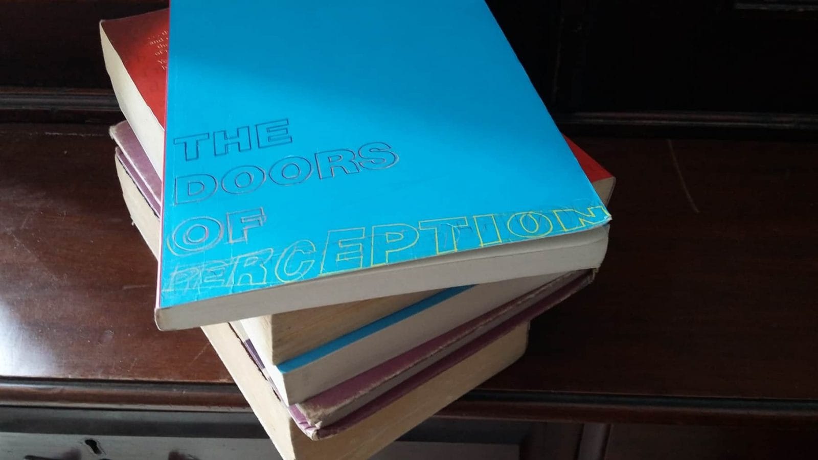 A pile of books sits on a wooden dressing table. The top book is titled 'The Doors Of Perception'