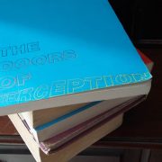 A pile of books sits on a wooden dressing table. The top book is titled 'The Doors Of Perception'