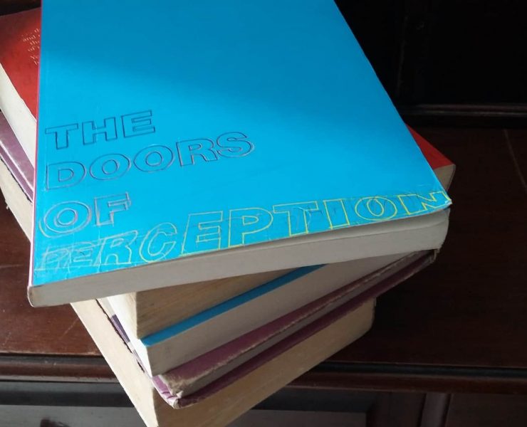 A pile of books sits on a wooden dressing table. The top book is titled 'The Doors Of Perception'
