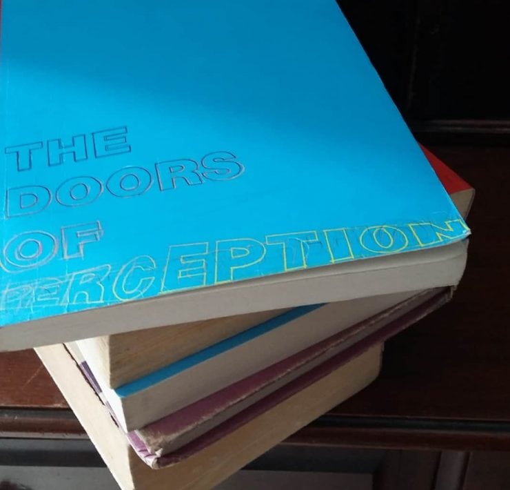 A pile of books sits on a wooden dressing table. The top book is titled 'The Doors Of Perception'