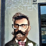 Graffiti picture of a hipster on a grey wall. The hipster has a quiff, glasses and a beard and twirled moustache. He wears a tweed suit with a bowtie