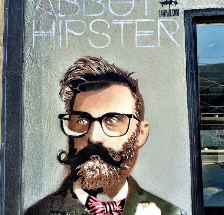 Graffiti picture of a hipster on a grey wall. The hipster has a quiff, glasses and a beard and twirled moustache. He wears a tweed suit with a bowtie
