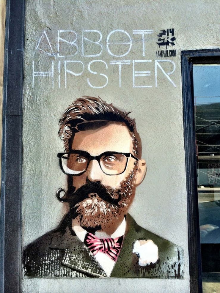 Graffiti picture of a hipster on a grey wall. The hipster has a quiff, glasses and a beard and twirled moustache. He wears a tweed suit with a bowtie