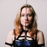 British Wrestler Pollyanna stands holding a sword betwwen her eyes as she stares into camera. She is wearing a costume reminiscent of a Game Of Thrones character