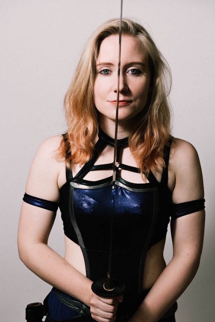 British Wrestler Pollyanna stands holding a sword betwwen her eyes as she stares into camera. She is wearing a costume reminiscent of a Game Of Thrones character