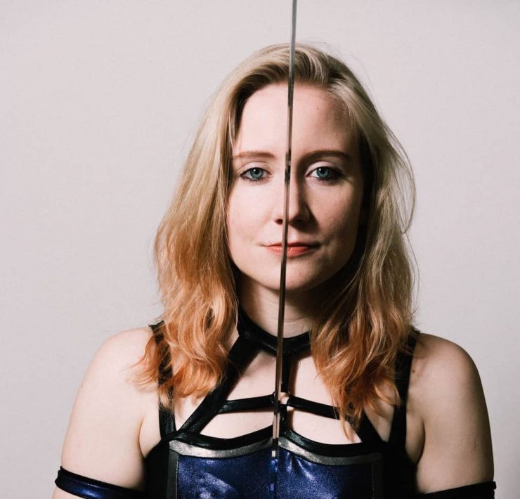 British Wrestler Pollyanna stands holding a sword betwwen her eyes as she stares into camera. She is wearing a costume reminiscent of a Game Of Thrones character