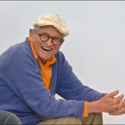 David Hockney sat in a chair with a white flat cap on with his hands claspt an leaning on his knees