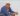 David Hockney sat in a chair with a white flat cap on with his hands claspt an leaning on his knees