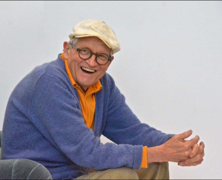 David Hockney sat in a chair with a white flat cap on with his hands claspt an leaning on his knees