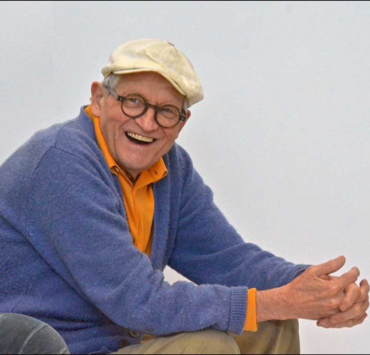 David Hockney sat in a chair with a white flat cap on with his hands claspt an leaning on his knees