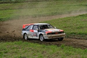 A white, red and black Audi Quattro raleigh car speeds through a mud track kicking up dirt as it goes 