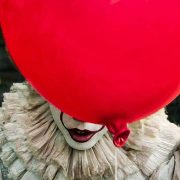 Picture of clown Pennywise from remake of IT. He is hiding behind red balloon so you can just see his mouth