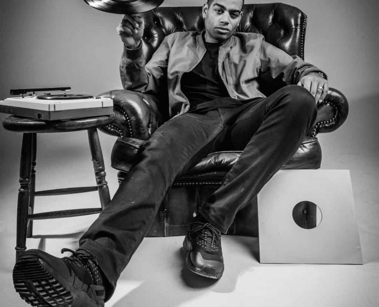 Comedian and rapper Doc Brown sits in a leather arm chair spinning a record on a finger of his right hand