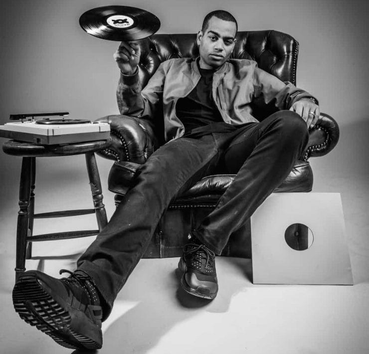 Comedian and rapper Doc Brown sits in a leather arm chair spinning a record on a finger of his right hand