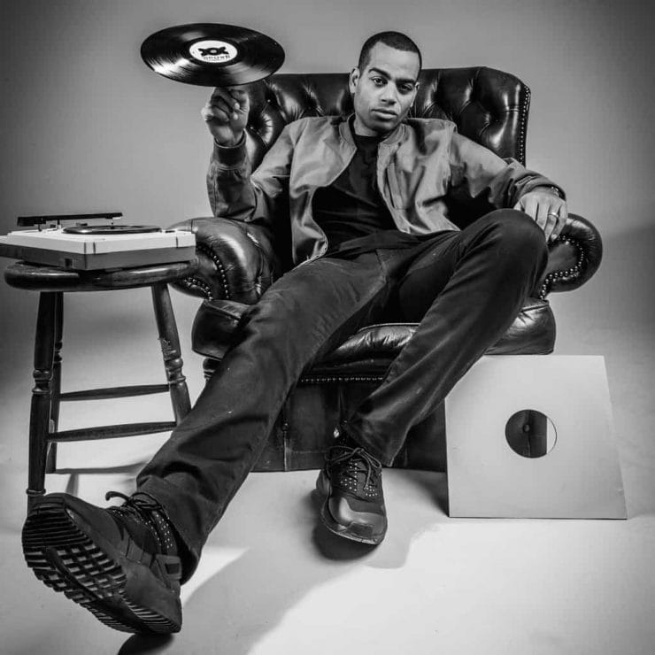 Comedian and rapper Doc Brown sits in a leather arm chair spinning a record on a finger of his right hand