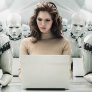 A women working on a laptop sits in a room full of human looking robots made out of shiny plastic