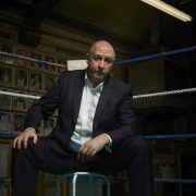 Boxing pundit Steve Bunce sits in the corner of a boxing ring on a stool looking moodily into camera like a gangster