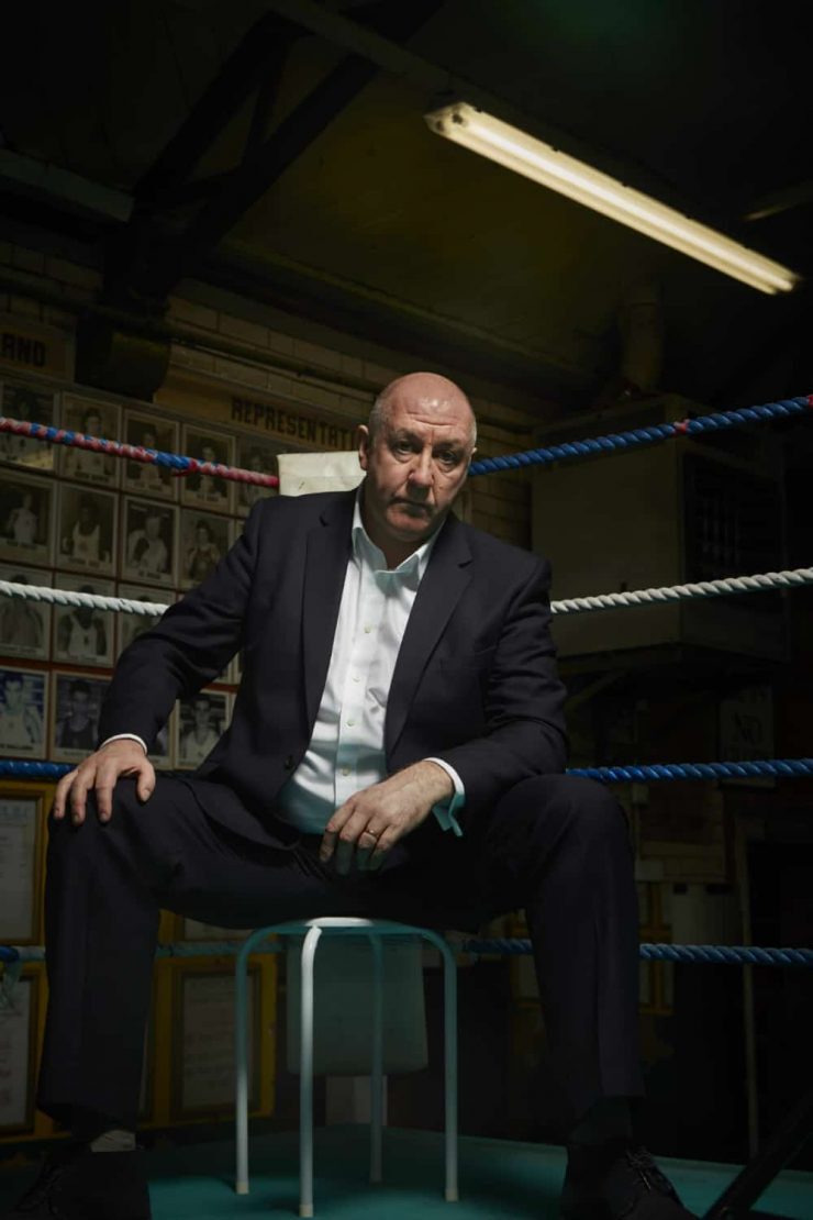 Boxing pundit Steve Bunce sits in the corner of a boxing ring on a stool looking moodily into camera like a gangster