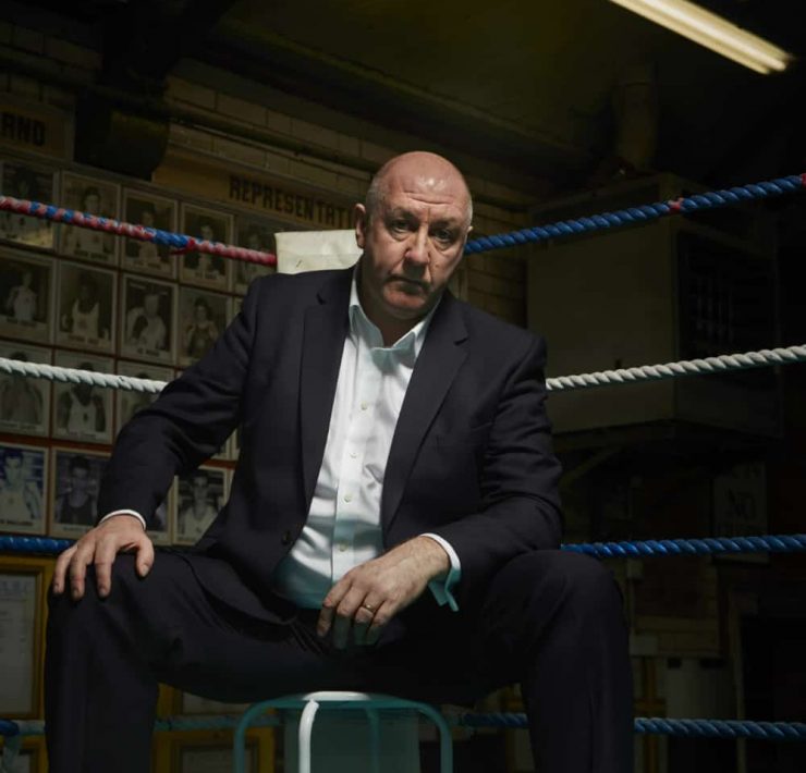Boxing pundit Steve Bunce sits in the corner of a boxing ring on a stool looking moodily into camera like a gangster