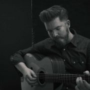 Folk musician John Smith stood playing his guitar with his eyes closed. The photograph is black and white and he is wering a dark shirt with white buttons
