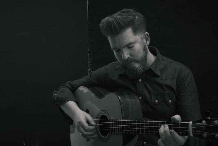 Folk musician John Smith stood playing his guitar with his eyes closed. The photograph is black and white and he is wering a dark shirt with white buttons