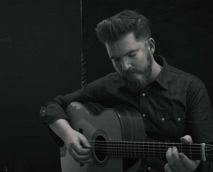 Folk musician John Smith stood playing his guitar with his eyes closed. The photograph is black and white and he is wering a dark shirt with white buttons