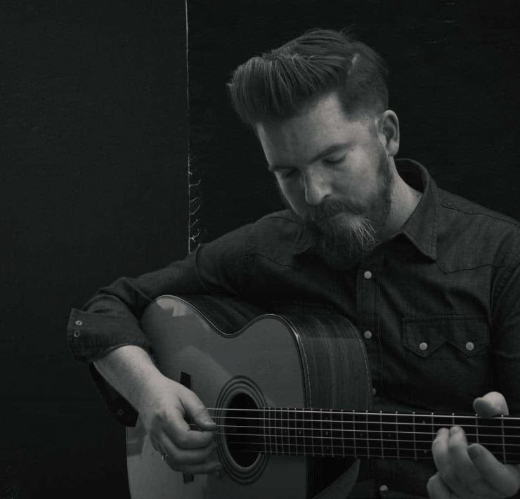 Folk musician John Smith stood playing his guitar with his eyes closed. The photograph is black and white and he is wering a dark shirt with white buttons