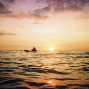 A surfer lies on his board ready to catch a wave as the sun sets below the horizon of the sea behind him
