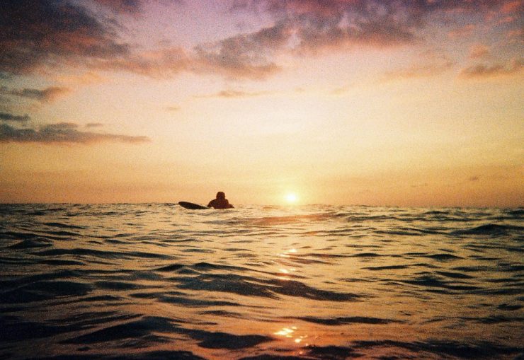 A surfer lies on his board ready to catch a wave as the sun sets below the horizon of the sea behind him