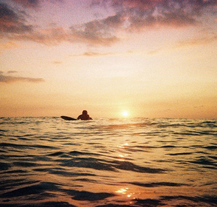 A surfer lies on his board ready to catch a wave as the sun sets below the horizon of the sea behind him