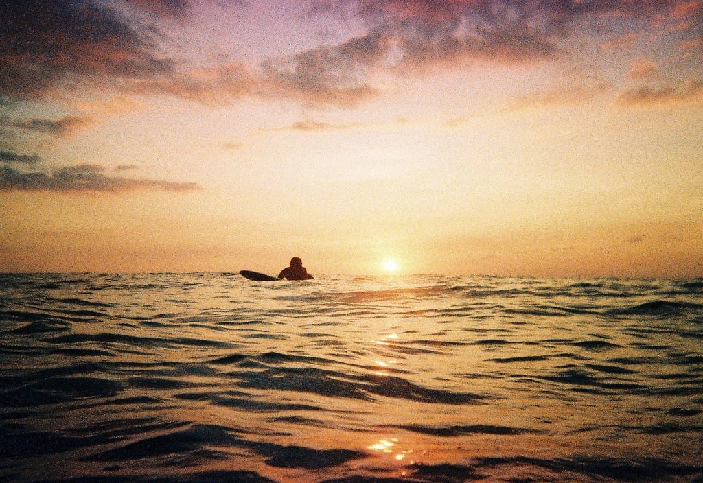 A surfer lies on his board ready to catch a wave as the sun sets below the horizon of the sea behind him