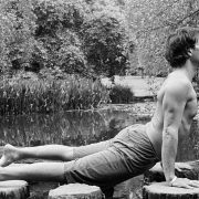 Wellness guru Chris James holds a prone cobra yoga move in a black and white picture. Chris is topless with shorts on. He's side on to camera with a backdrop of trees