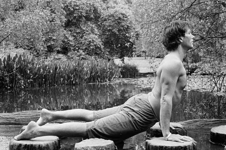 Wellness guru Chris James holds a prone cobra yoga move in a black and white picture. Chris is topless with shorts on. He's side on to camera with a backdrop of trees