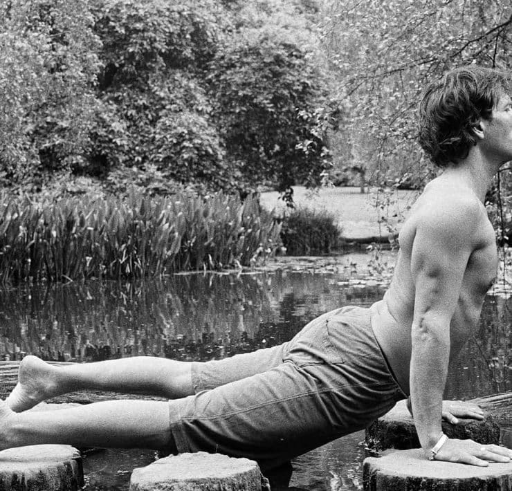 Wellness guru Chris James holds a prone cobra yoga move in a black and white picture. Chris is topless with shorts on. He's side on to camera with a backdrop of trees