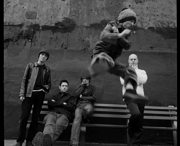 Radiohead stand and sit behind Thom Yorke as he does a flying kick to camera