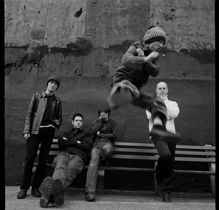 Radiohead stand and sit behind Thom Yorke as he does a flying kick to camera