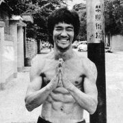 A shirtless Bruce Lee stands shirtless on a street corner, smiling with his hands clasped together