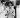 A shirtless Bruce Lee stands shirtless on a street corner, smiling with his hands clasped together