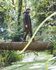 Aldo Kane crossing a log bridge in the woods wearing Fjӓllrӓven's sustainable clothing range