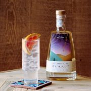 A bottle of El Rayo tequilla on a table with a glass of tequilla and tonic next to it