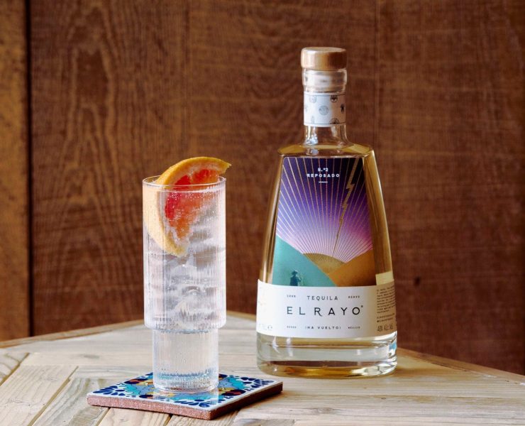 A bottle of El Rayo tequilla on a table with a glass of tequilla and tonic next to it