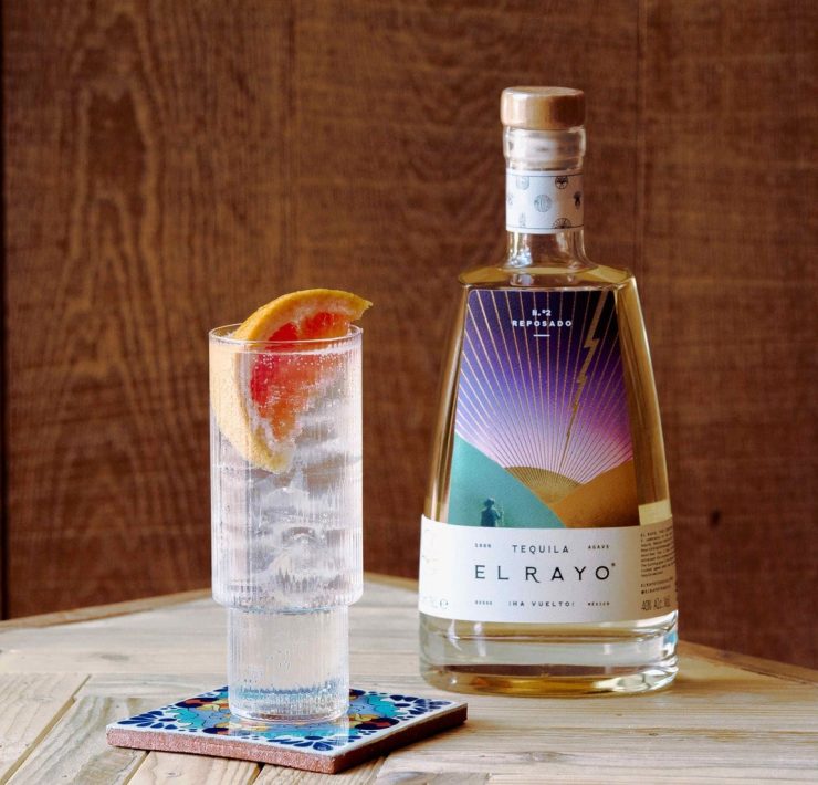 A bottle of El Rayo tequilla on a table with a glass of tequilla and tonic next to it