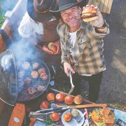 Christian Stevenson aka DJ BBQ holds up a burger to camera while BBQing