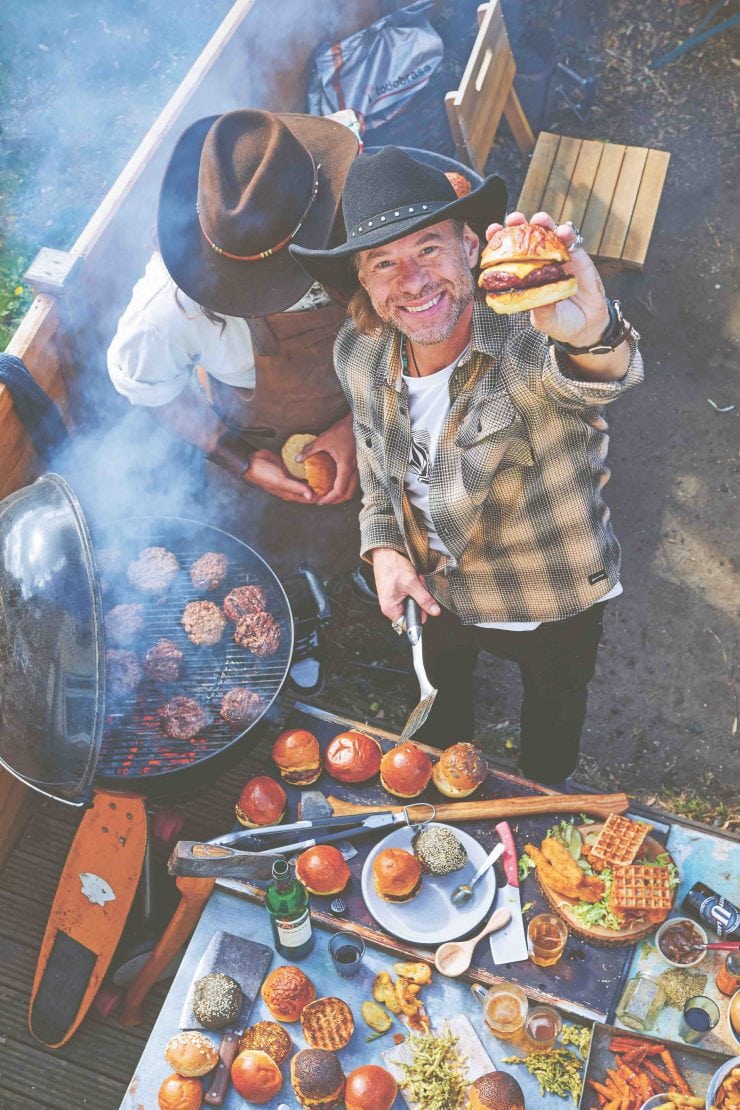 Christian Stevenson aka DJ BBQ holds up a burger to camera while BBQing