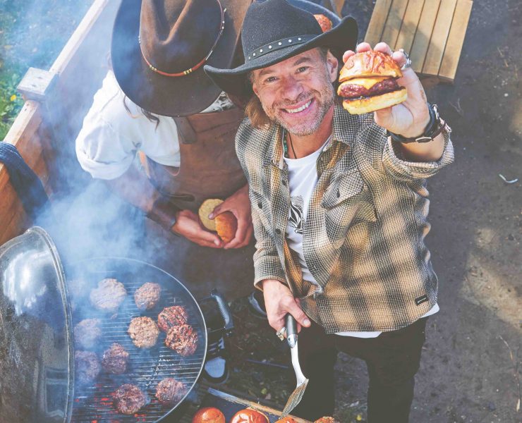 Christian Stevenson aka DJ BBQ holds up a burger to camera while BBQing