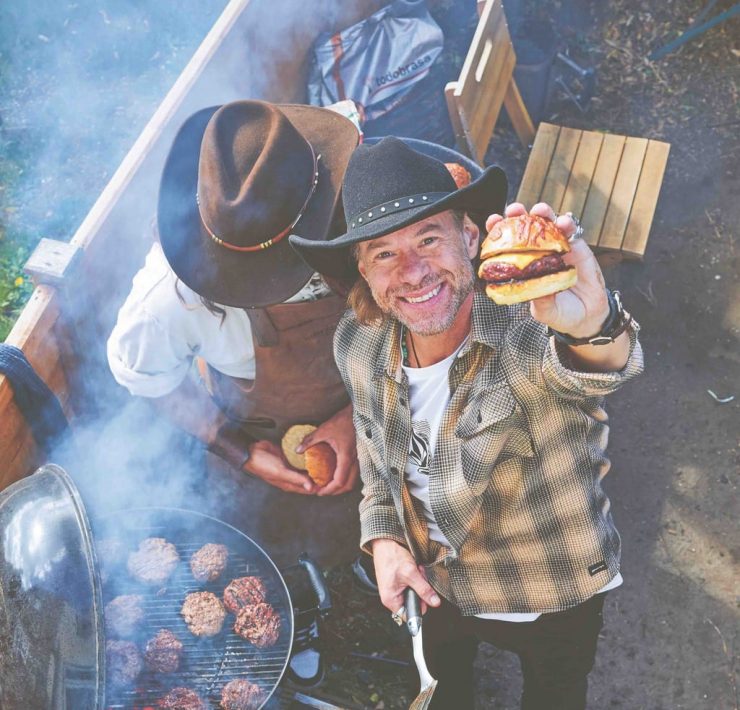 Christian Stevenson aka DJ BBQ holds up a burger to camera while BBQing