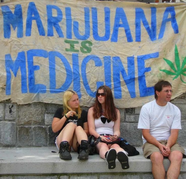 Two girls and a man sit in front of a sign saying 'Marijuana is Medicine' at Vancouver Hemp Rally