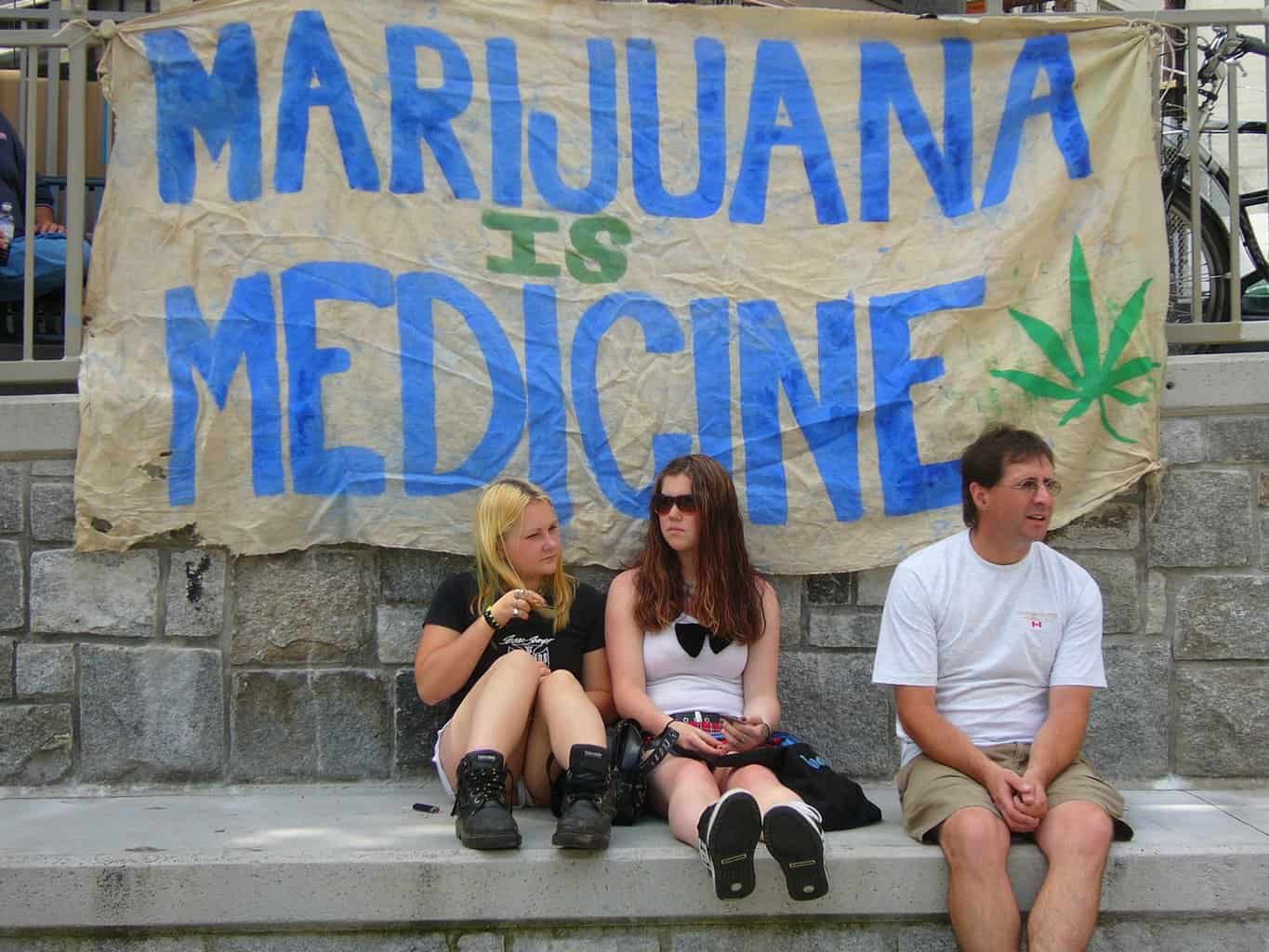 Two girls and a man sit in front of a sign saying 'Marijuana is Medicine' at Vancouver Hemp Rally